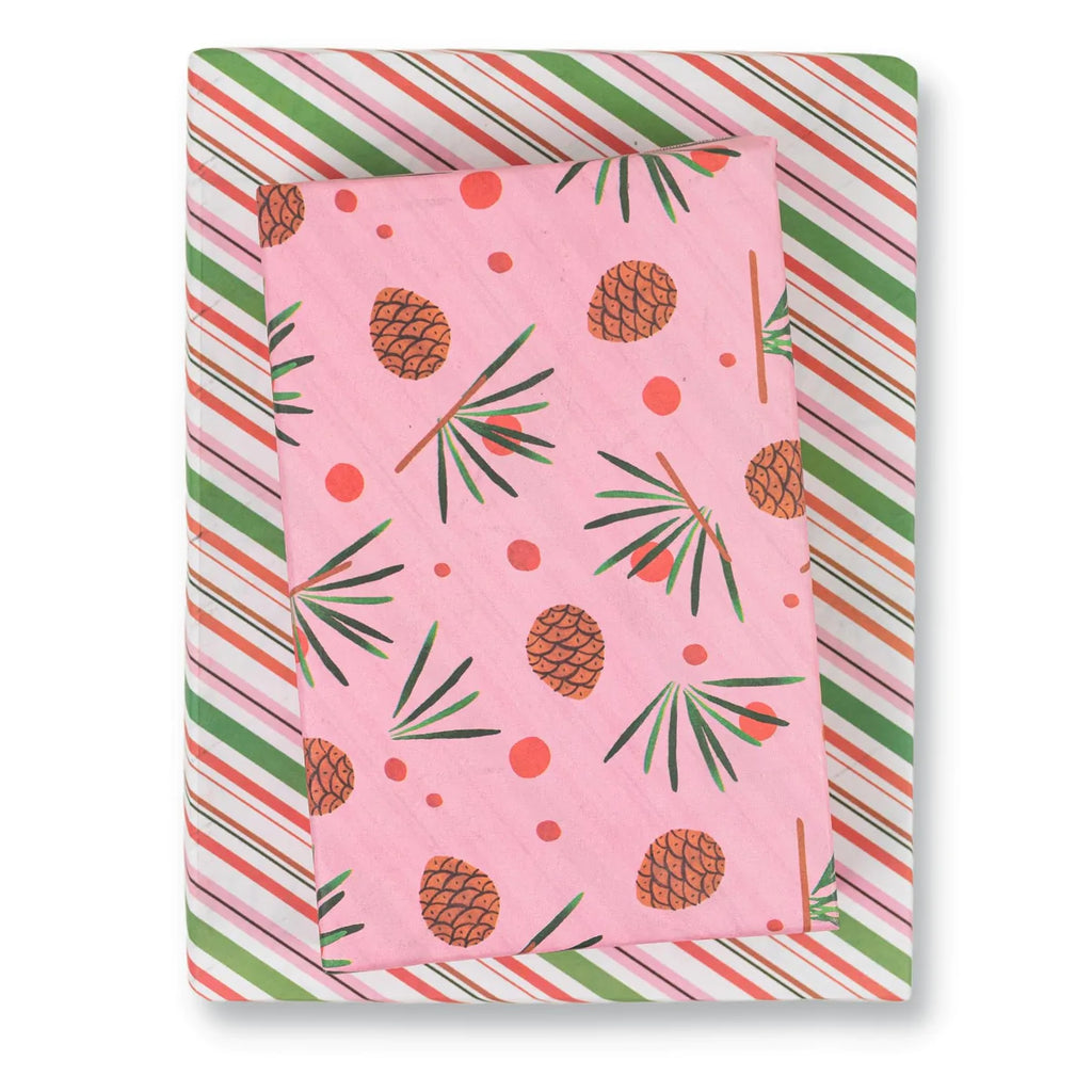 An image of a package of Misha Zadeh for Wrappily Pinecone gift wrap: watercolor-painted Pinecones and pine needles on a pink background, with a snippet of the backside print, which is diagonal stripes in pinks, reds, and greens, mimicking candy cane striping.