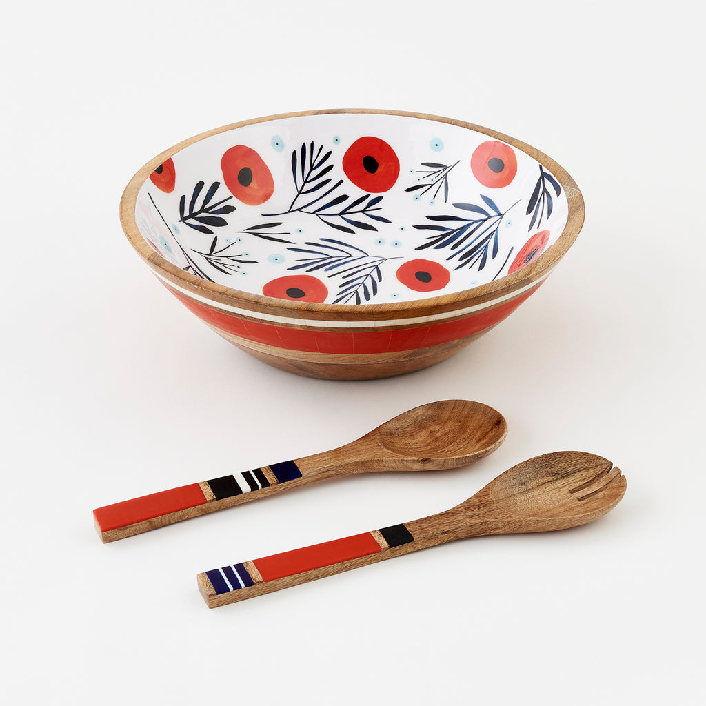 Photo of a Mango Wood bowl with Red and Navy Watercolor Poppies on a field of white, with light blue berries interspersed throughout. Bowl has a large red stripe and a small white stripe handpainted on the exterior. To the side are a pair of coordinating, striped wooden tongs. Designed by Misha Zadeh for 180 Degrees