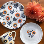 Flatlay of Misha Zadeh Poppy Love 9", 7.5" and ceramic mugs, along with a couple orange dahlias arranged near them