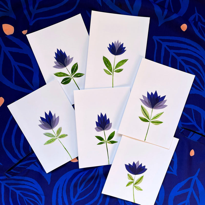 A set of six handmade, cut-paper greeting cards by Seattle artist Misha Zadeh sit atop a background of "skeleton Leaves" print. The cards are one of a kind and feature deep purple and lavender, overlapping translucent, colorful vellumpetals, with green leaves that are made of hand-painted card stock.