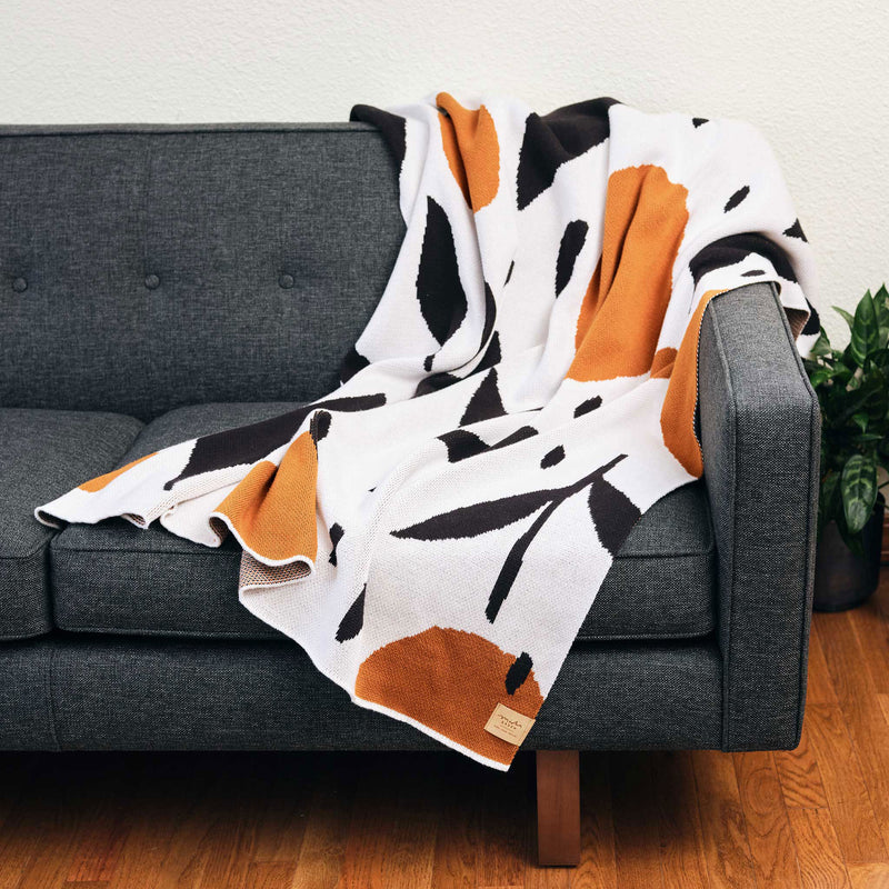 Misha Zadeh Rain Garden Throw Blanket in Black, White, and Copper, loosely draped over a modern charcoal gray upholstery couch.