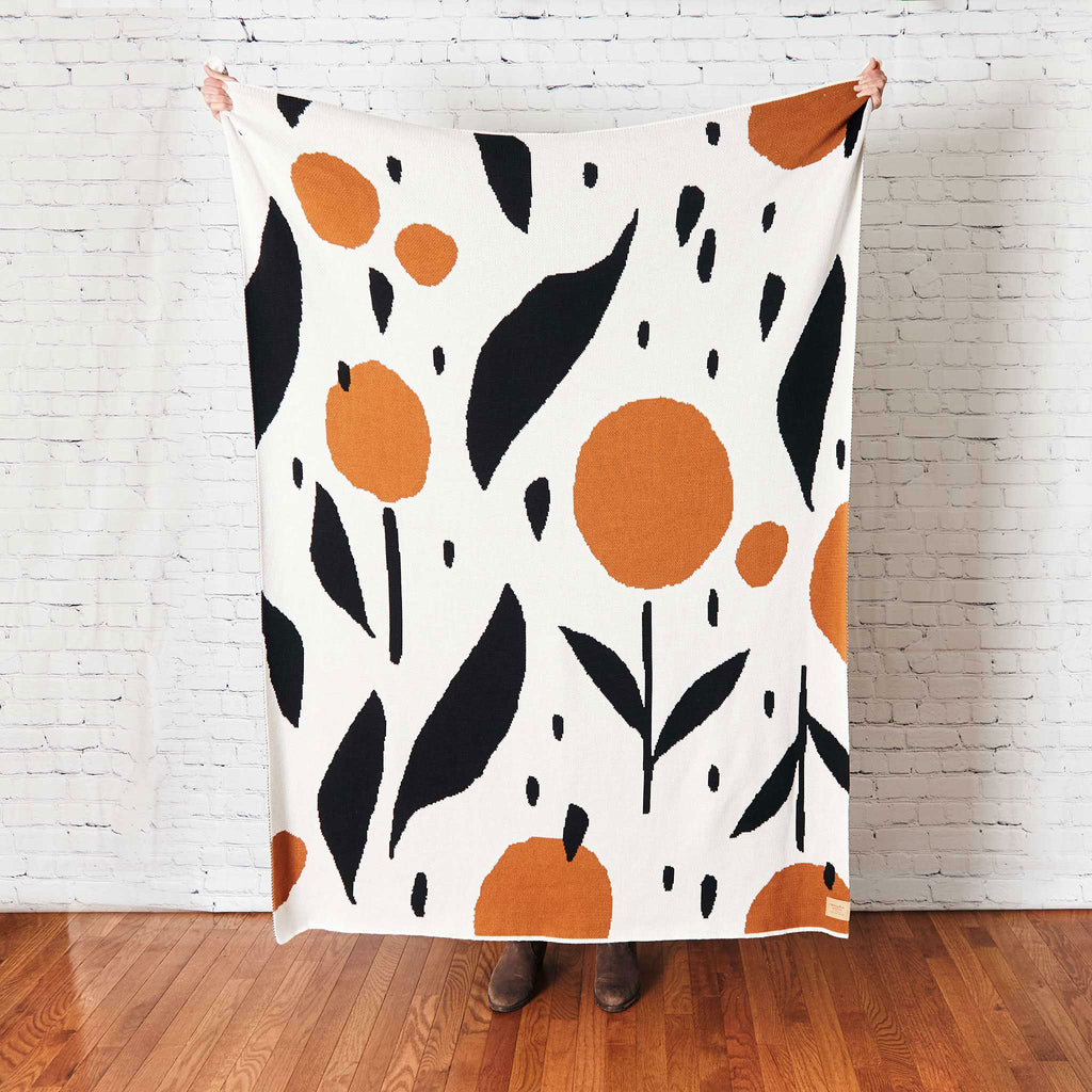 Misha Zadeh Rain Garden Knit Blanket in Black, White, and Copper, held up by a person standing in front of a white brick wall.