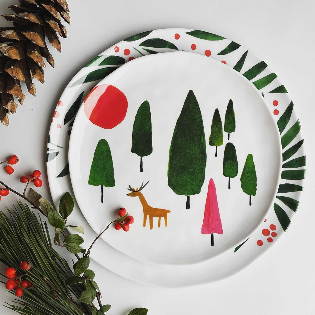 Misha Zadeh Winter Forest Deer Melamine Plate layered on top of her Winter Berries platter. Styled on a white table with a pinecone and orange winter berries layered on top