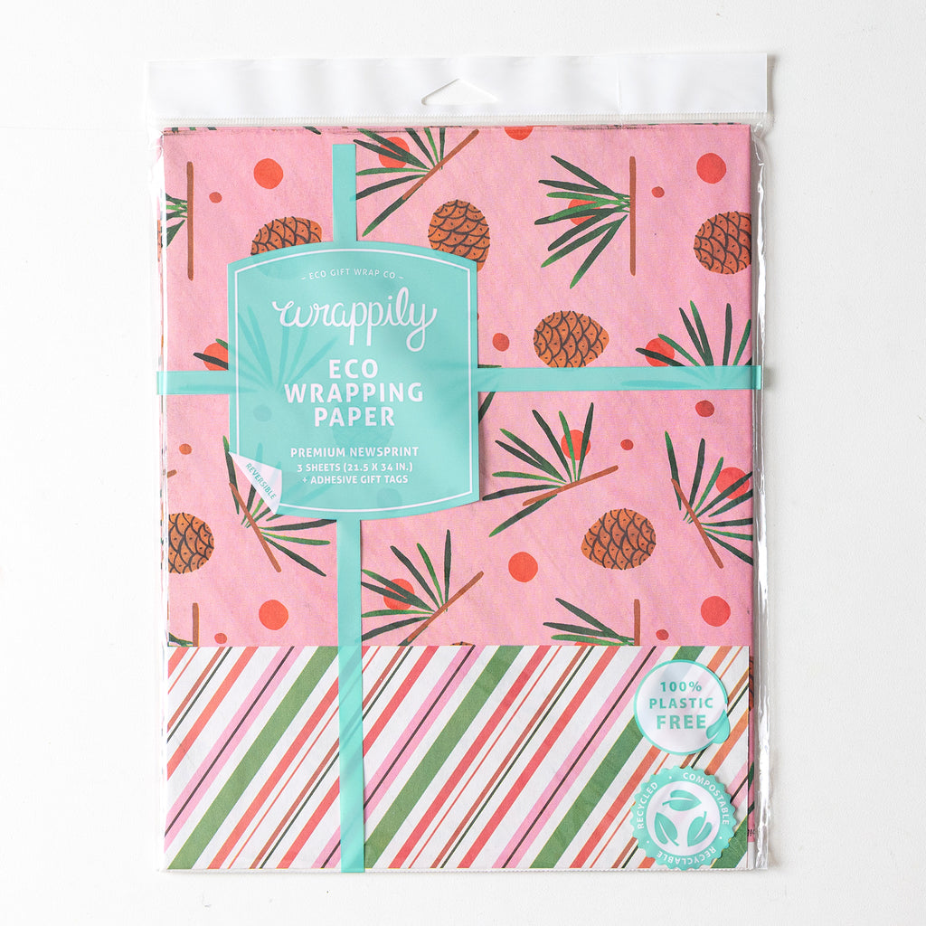An image of a package of Misha Zadeh for Wrappily Pinecone gift wrap: watercolor-painted Pinecones and pine needles on a pink background, with a snippet of the backside print, which is diagonal stripes in pinks, reds, and greens, mimicking candy cane striping.