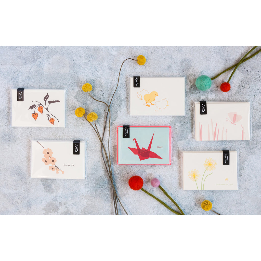 Collection of Misha Zadeh for Turquoise Creative Letterpress note cards, styled on a concrete background with craspedia and felted pom pom flowers. Featured cards are Chinese Lanterns, Pink Dogwoods, Baby Chick, Dandelion, Pink butterfly and Red Crane