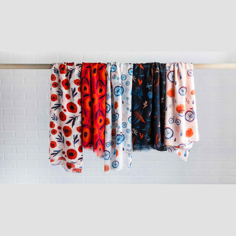 Misha Zadeh Modal Scarves hanging from a rod. Poppy Specimen, Country Poppies in Red, Misty Bicycle in White, Under The Sea in Black, and Misty Bicycle in Pink