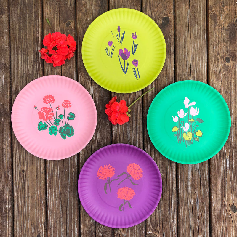 Les Fleurs, Floral Melamine Plate Sets by Misha Zadeh, featuring brush pen artwork of geraniums, mums, cyclamen, and crocus flowers. Styled atop dark wood decking with two real, coral geraniums placed around the plates.
