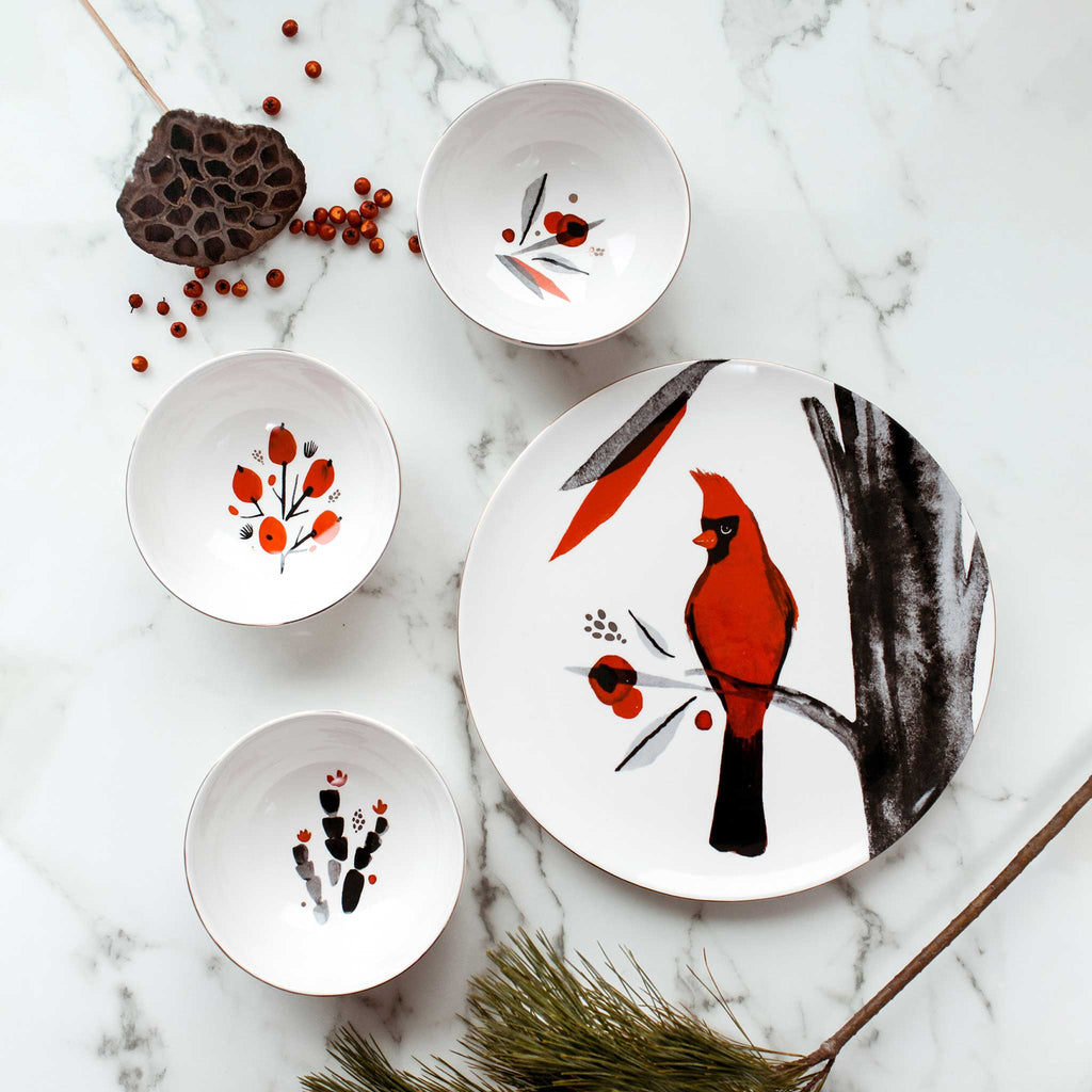 Misha Zadeh Cardinal And Berries Ceramic Plate and Bowl collection, on a marble table top with winter foliage and berries laid out around them